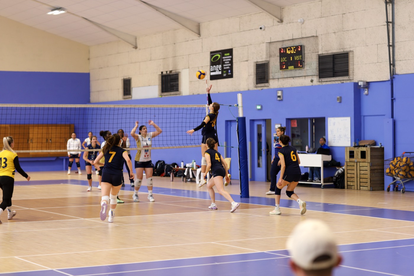 Phase de jeu au filet dans un gymnase, joueuses en maillots bleu nuit et jaune