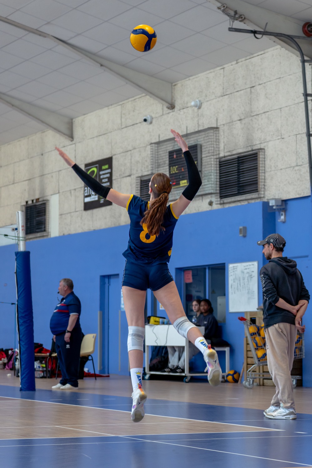 Une joueuse en maillot bleu nuit bondit, bras tendus, pour servir pendant un match